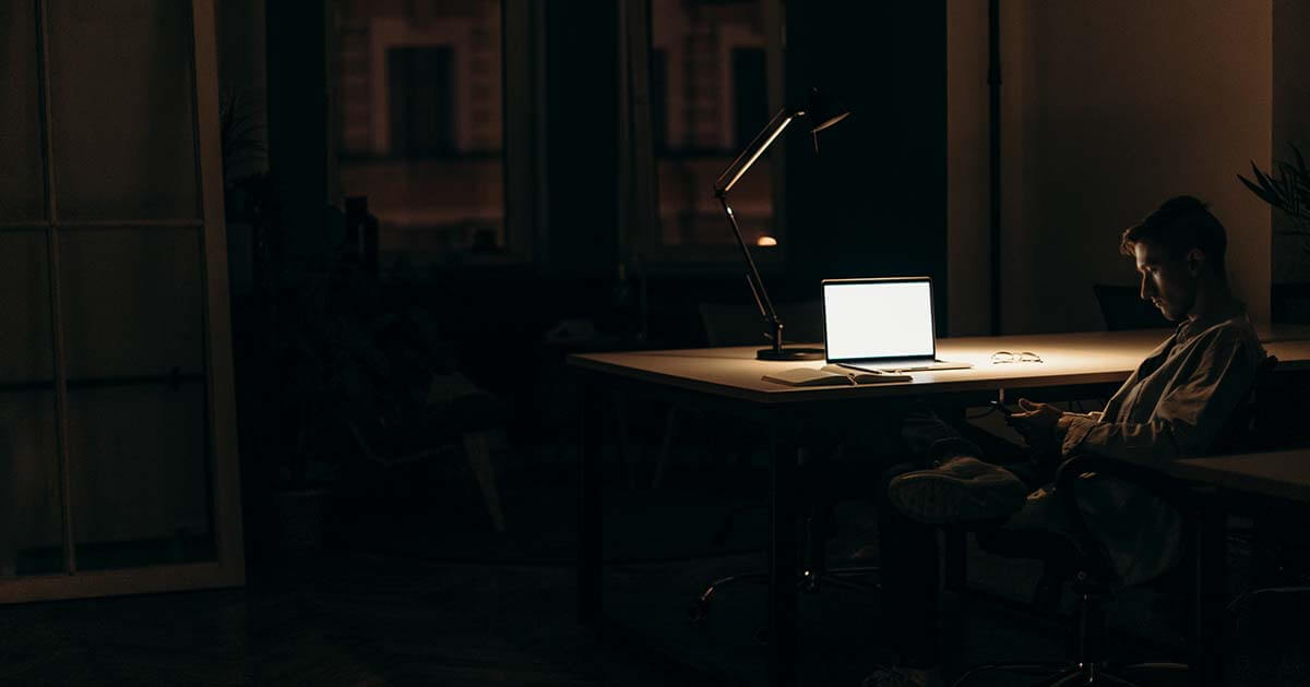 A man sitting at an office desk in the dark browsing his phone with a laptop open